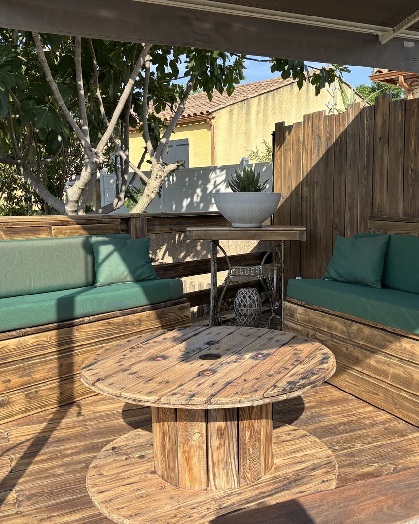 Salon de jardin sur mesure en bois : deux banquettes avec coussins verts et table basse bobine, sur une terrasse bois réalisée par Articréa